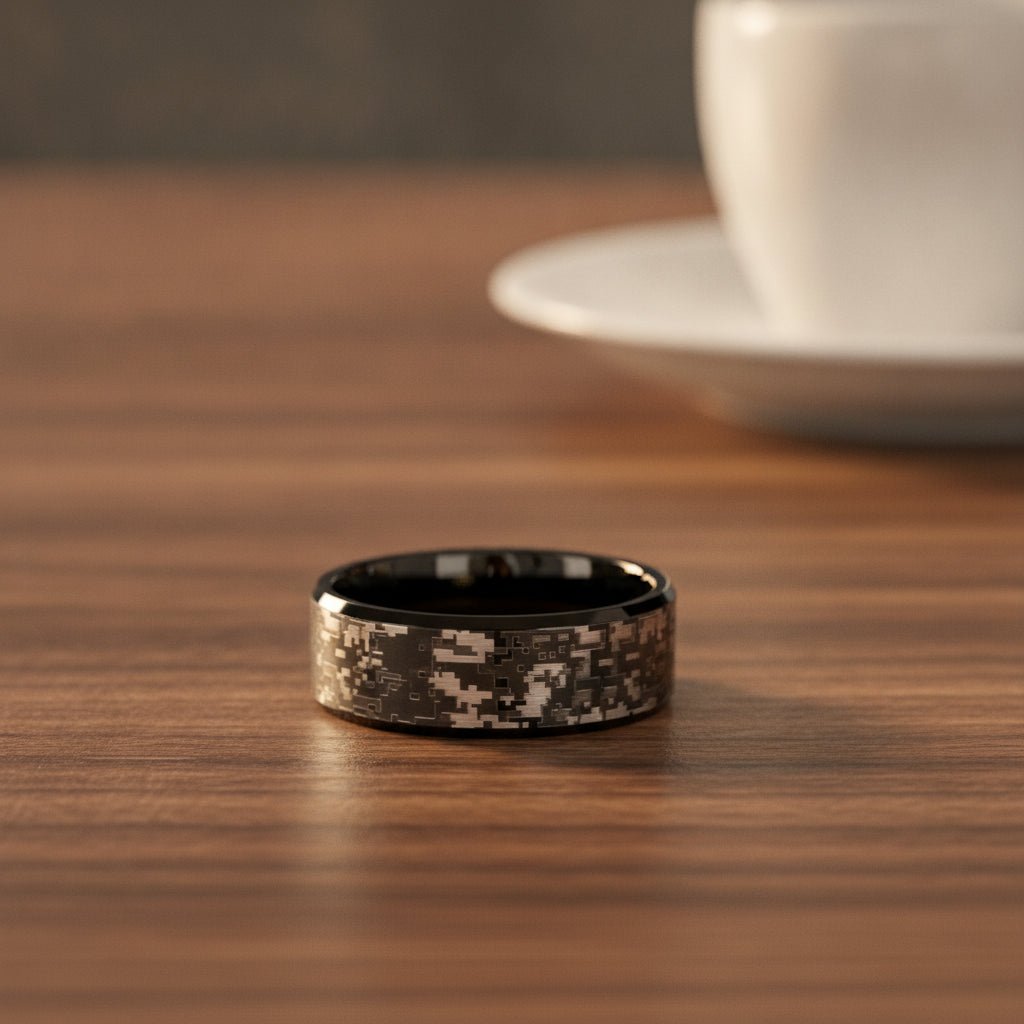 SMOKESCREEN black tungsten carbide wedding band with engraved digital camouflage pattern running around the band and polished edges resting on a walnut wood counter, lifestyle scene