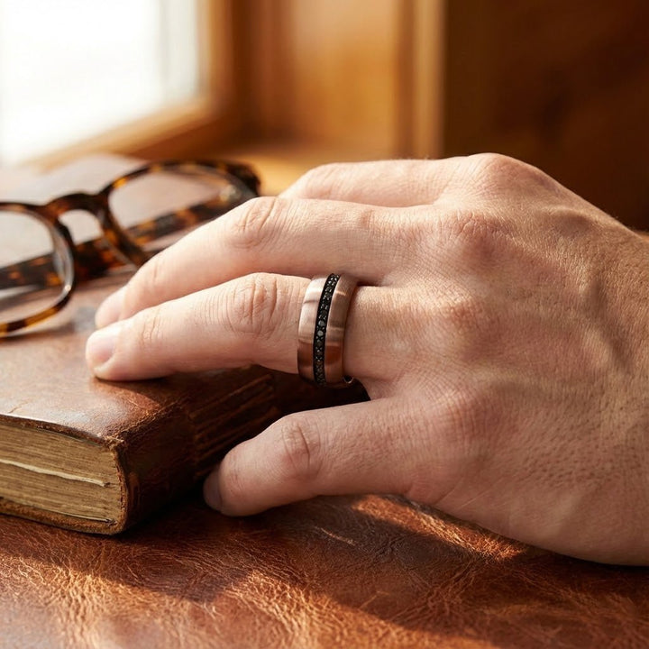 Aydins Jewelry Modern men’s tungsten wedding band with espresso finish and black sapphires on a mans ring finger near a book