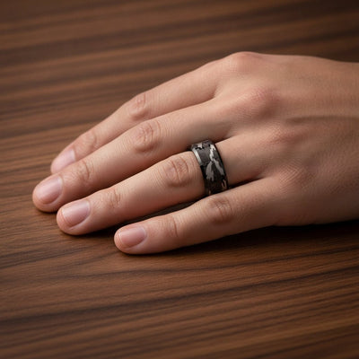 CONQUEST beveled black tungsten carbide wedding band with black and gray camouflage pattern inlay running around the band and polished edges worn on man's hand resting on walnut wood, lifestyle photo