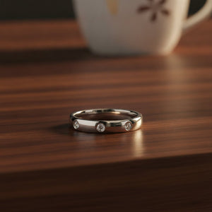 ISABELLA domed white tungsten wedding band with three small diamond accent stones set in a row on the center of the band and polished edges resting on a walnut wood counter, lifestyle scene