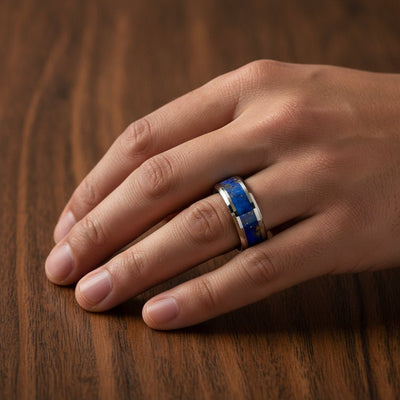 OSIAS polished silver tungsten wedding band with blue lapis stone inlay running around the band and polished beveled edges worn on man's hand resting on walnut wood, lifestyle photo