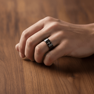 SANCTUS black tungsten wedding band with minimalist cross engraving worn on man's hand resting on walnut table, lifestyle photo