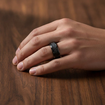 VESUVIUS polished black ceramic wedding band with black and gray lava rock stone inlay running around the band and polished beveled edges worn on man's hand resting on walnut wood, lifestyle photo